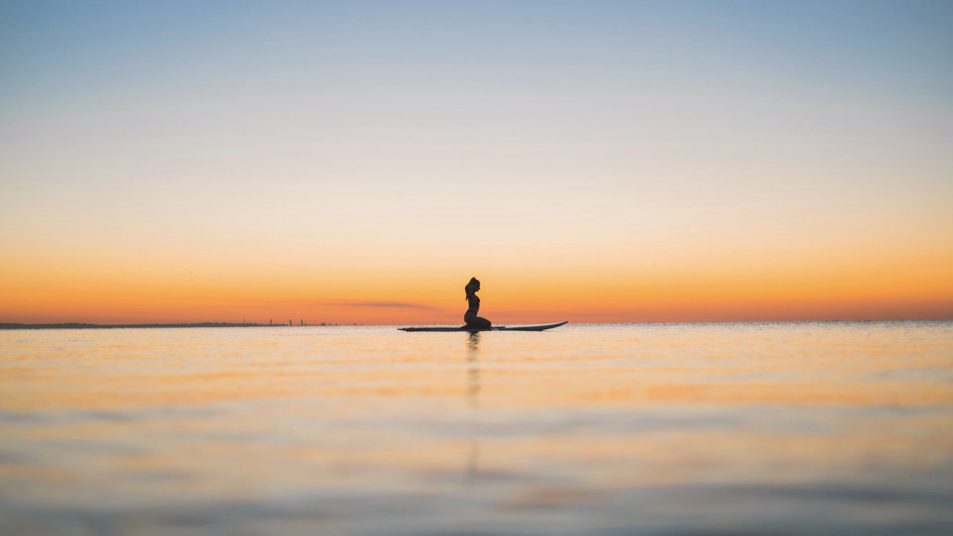 A woman sitting still on a paddleboard on calm open water at dusk